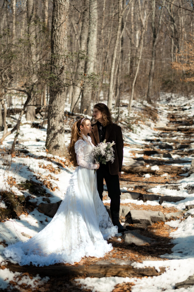 Cinematic photograph of an adventurous couple eloping in the forest, featuring the bride in a lace vintage wedding dress holding a floral bouquet while the groom is in a rustic suit. Captured by a destination wedding photographer in a documentary style.