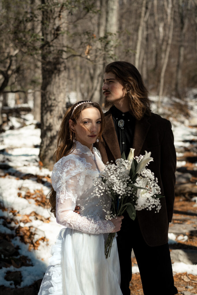 Cinematic photograph of an adventurous couple eloping in the forest, featuring the bride in a lace vintage wedding dress holding a floral bouquet while the groom is in a rustic suit. Captured by a destination wedding photographer in a documentary style.