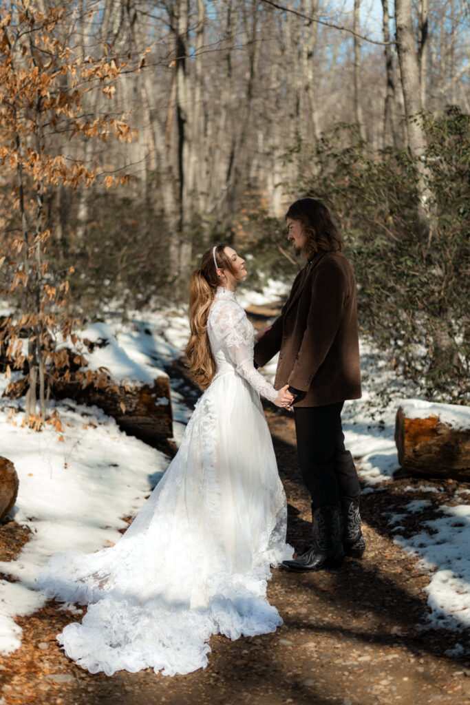 Cinematic photograph of an adventurous couple eloping in the forest, featuring the bride in a lace vintage wedding dress holding a floral bouquet while the groom is in a rustic suit. Captured by a destination wedding photographer in a documentary style.