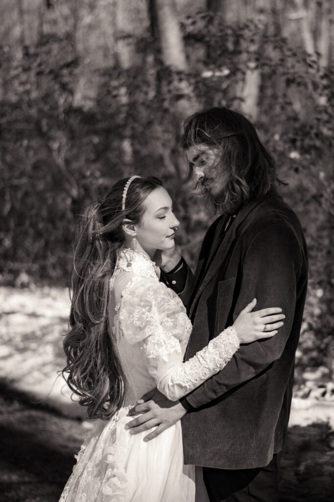 Cinematic photograph of an adventurous couple eloping in the forest, featuring the bride in a lace vintage wedding dress holding a floral bouquet while the groom is in a rustic suit. Captured by a destination wedding photographer in a documentary style.