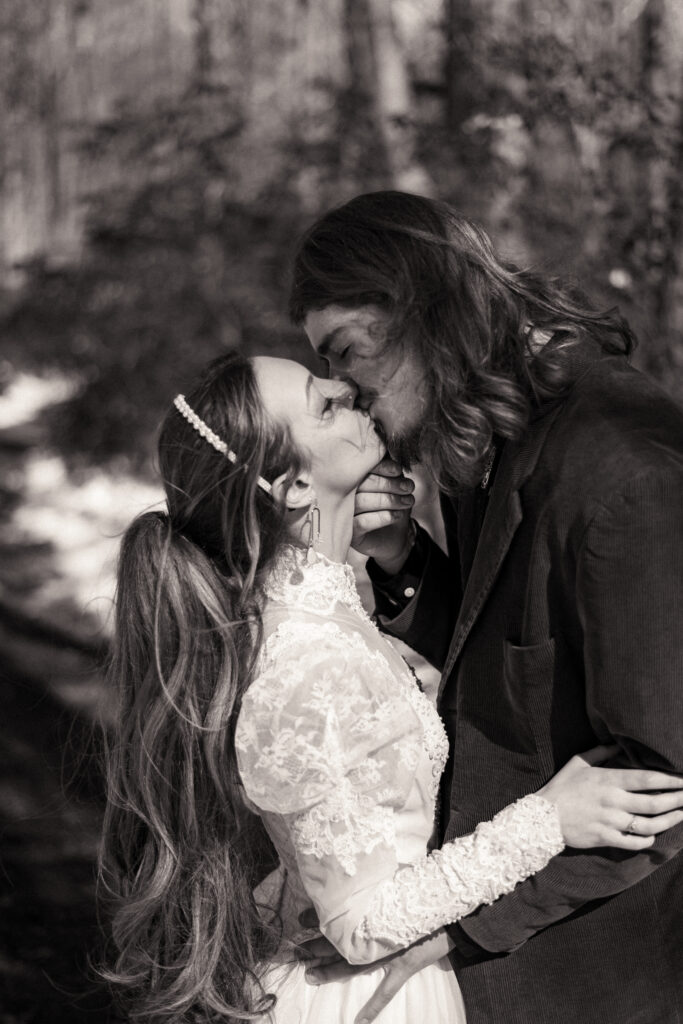 Cinematic photograph of an adventurous couple eloping in the forest, featuring the bride in a lace vintage wedding dress holding a floral bouquet while the groom is in a rustic suit. Captured by a destination wedding photographer in a documentary style.