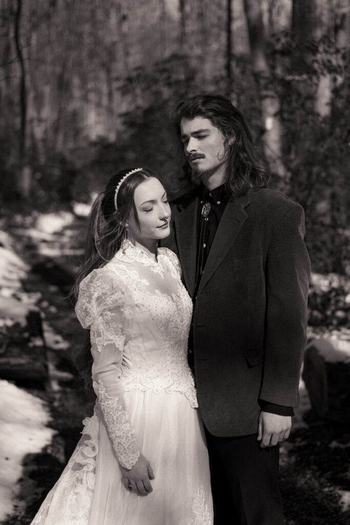 Cinematic photograph of an adventurous couple eloping in the forest, featuring the bride in a lace vintage wedding dress holding a floral bouquet while the groom is in a rustic suit. Captured by a destination wedding photographer in a documentary style.