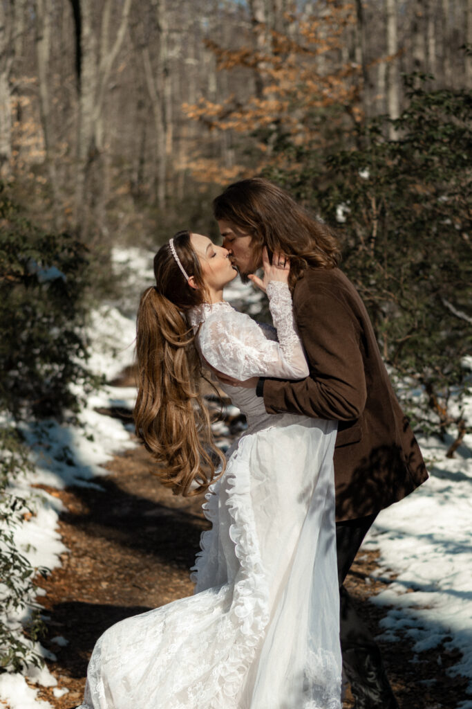 Cinematic photograph of an adventurous couple eloping in the forest, featuring the bride in a lace vintage wedding dress holding a floral bouquet while the groom is in a rustic suit. Captured by a destination wedding photographer in a documentary style.