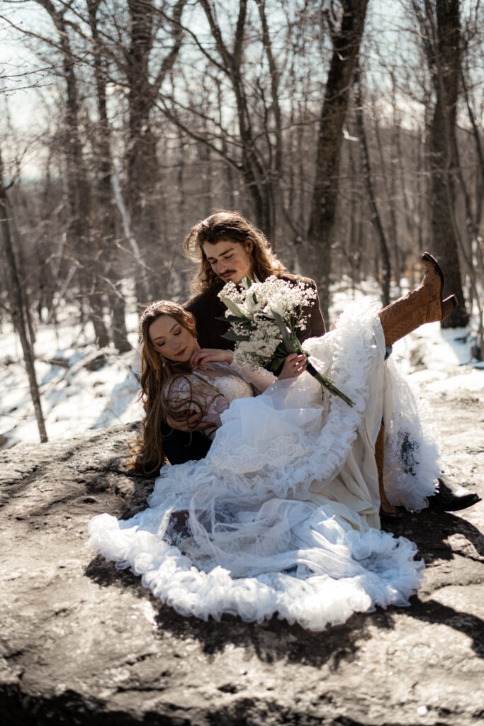 Cinematic photograph of an adventurous couple eloping in the forest, featuring the bride in a lace vintage wedding dress holding a floral bouquet while the groom is in a rustic suit. Captured by a destination wedding photographer in a documentary style.