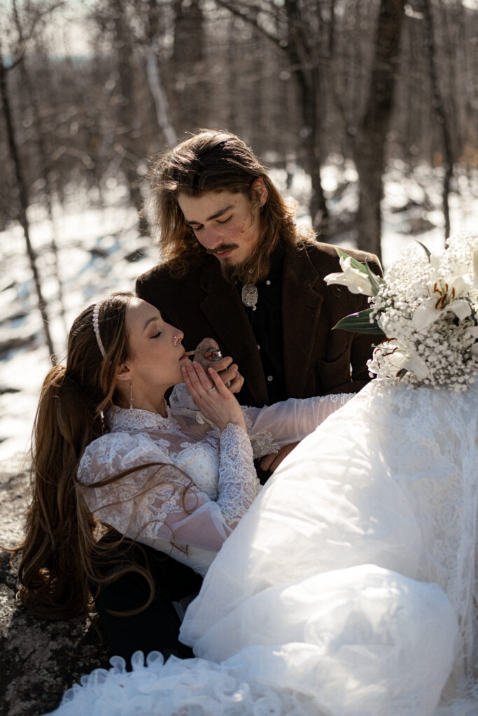 Cinematic photograph of an adventurous couple eloping in the forest, featuring the bride in a lace vintage wedding dress holding a floral bouquet while the groom is in a rustic suit. Captured by a destination wedding photographer in a documentary style.