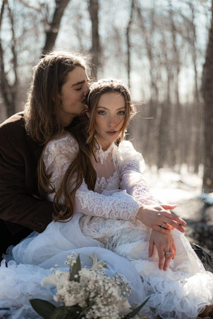 Cinematic photograph of an adventurous couple eloping in the forest, featuring the bride in a lace vintage wedding dress holding a floral bouquet while the groom is in a rustic suit. Captured by a destination wedding photographer in a documentary style.