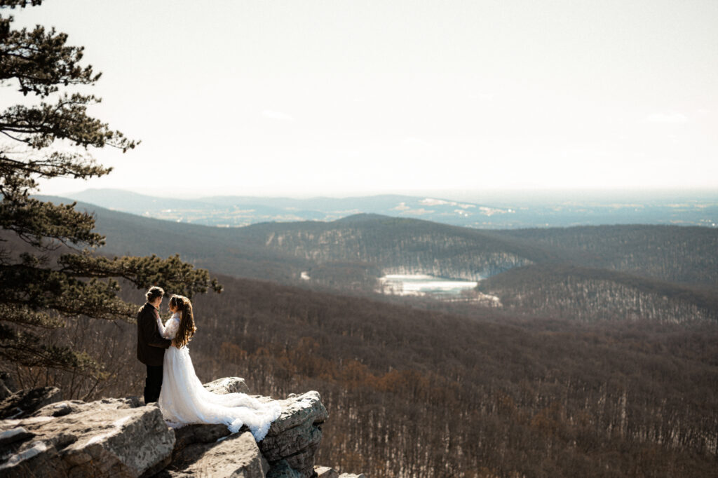 Cinematic photograph of an adventurous couple eloping in the forest, featuring the bride in a lace vintage wedding dress holding a floral bouquet while the groom is in a rustic suit. Captured by a destination wedding photographer in a documentary style.