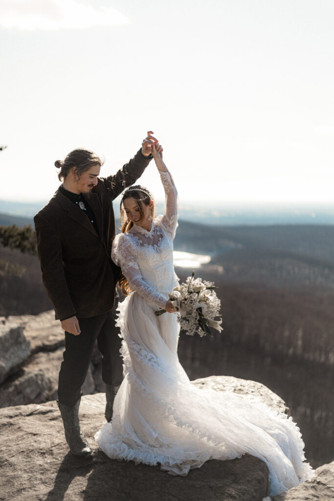 Cinematic photograph of an adventurous couple eloping in the forest, featuring the bride in a lace vintage wedding dress holding a floral bouquet while the groom is in a rustic suit. Captured by a destination wedding photographer in a documentary style.