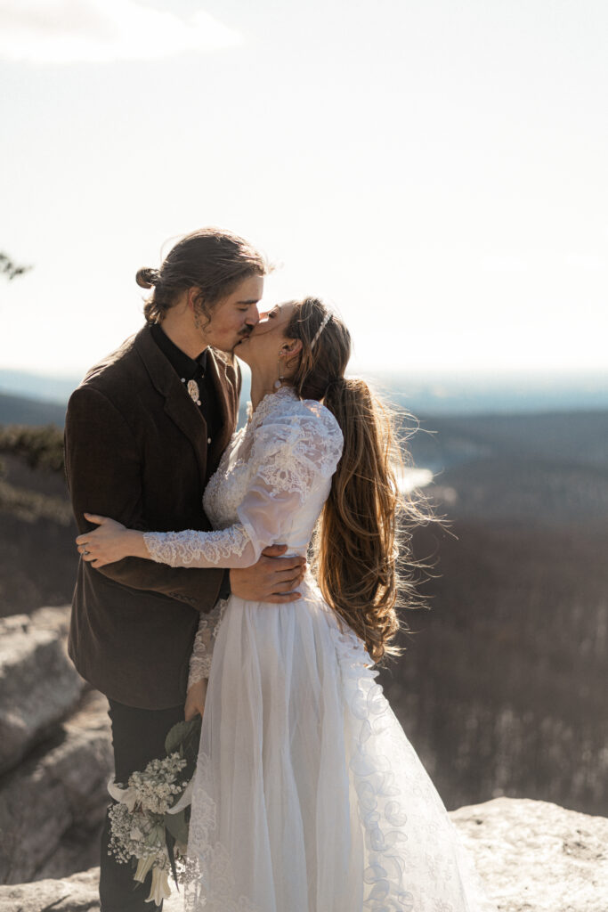 Cinematic photograph of an adventurous couple eloping in the forest, featuring the bride in a lace vintage wedding dress holding a floral bouquet while the groom is in a rustic suit. Captured by a destination wedding photographer in a documentary style.