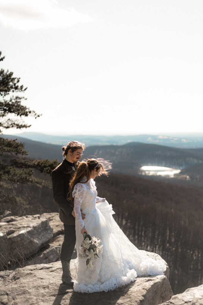 Cinematic photograph of an adventurous couple eloping in the forest, featuring the bride in a lace vintage wedding dress holding a floral bouquet while the groom is in a rustic suit. Captured by a destination wedding photographer in a documentary style.