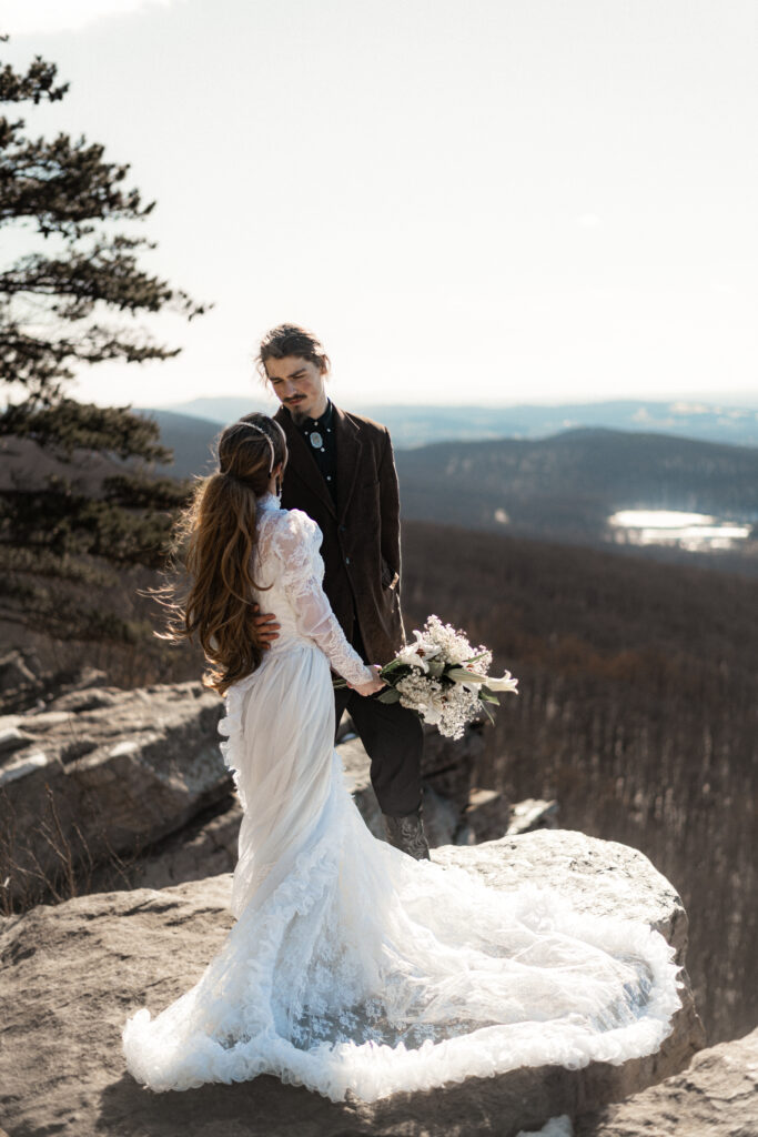 Cinematic photograph of an adventurous couple eloping in the forest, featuring the bride in a lace vintage wedding dress holding a floral bouquet while the groom is in a rustic suit. Captured by a destination wedding photographer in a documentary style.