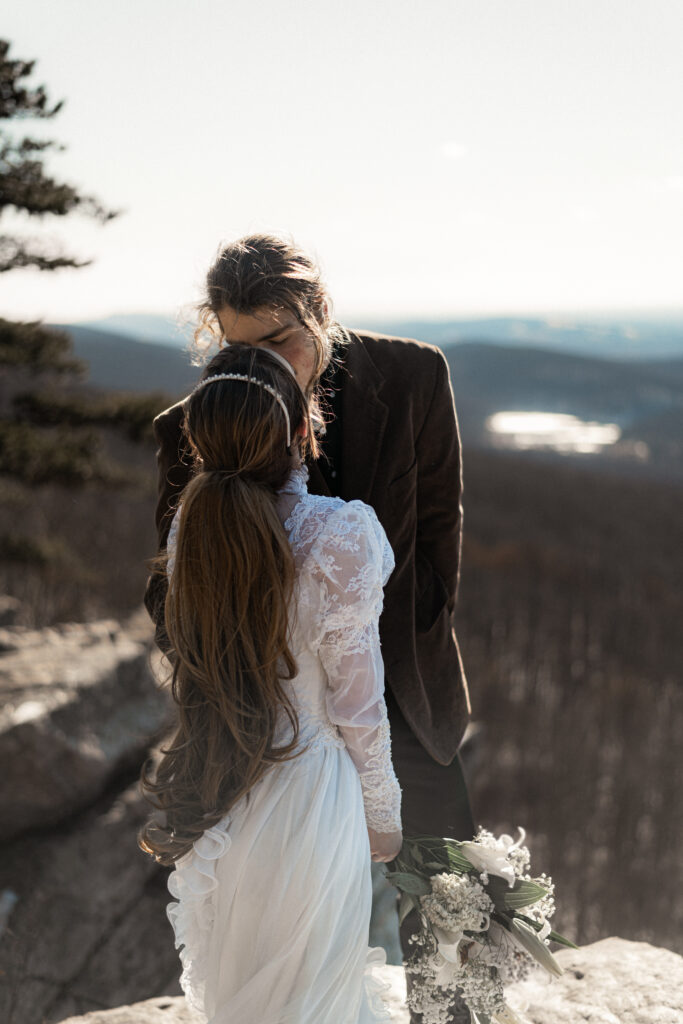 Cinematic photograph of an adventurous couple eloping in the forest, featuring the bride in a lace vintage wedding dress holding a floral bouquet while the groom is in a rustic suit. Captured by a destination wedding photographer in a documentary style.