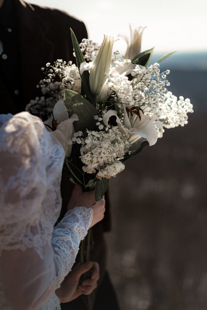 Cinematic photograph of an adventurous couple eloping in the forest, featuring the bride in a lace vintage wedding dress holding a floral bouquet while the groom is in a rustic suit. Captured by a destination wedding photographer in a documentary style.