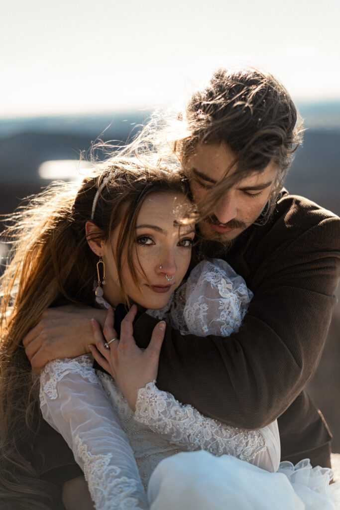 Cinematic photograph of an adventurous couple eloping in the forest, featuring the bride in a lace vintage wedding dress holding a floral bouquet while the groom is in a rustic suit. Captured by a destination wedding photographer in a documentary style.